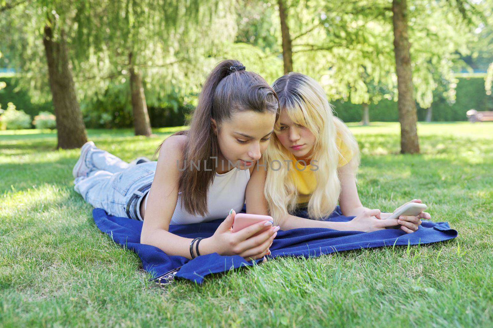 Two young female teenagers lying on grass in park with smartphones by VH-studio