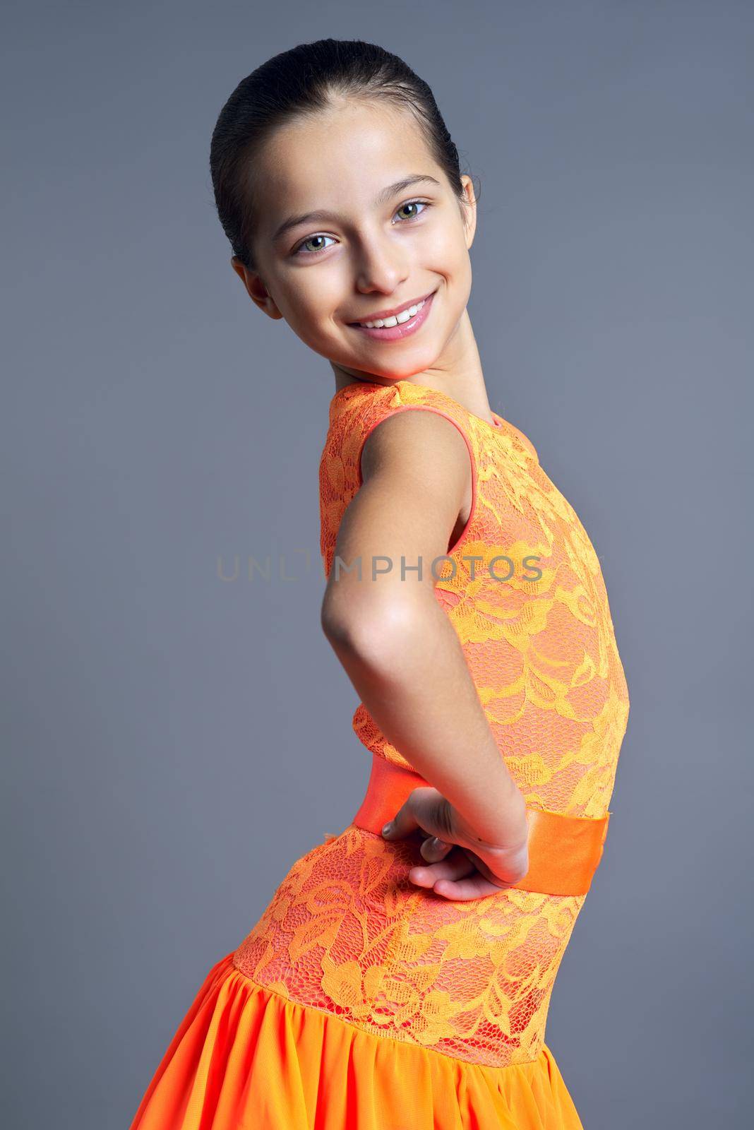 Portrait of smiling child girl in orange sports dress posing looking at camera on gray studio background, sport dances ballroom and latin american for children and teenagers