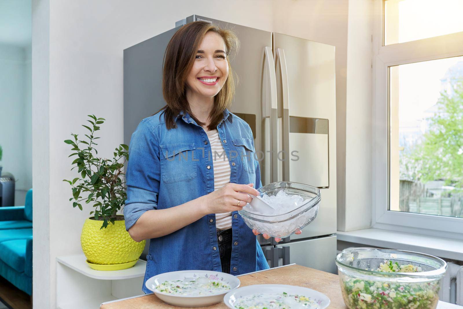 Smiling woman in kitchen near modern chrome large refrigerator with ice for cooling food, vegetable salad in bowl on table. Eating at home, lifestyle, household, dieting, healthy food concept