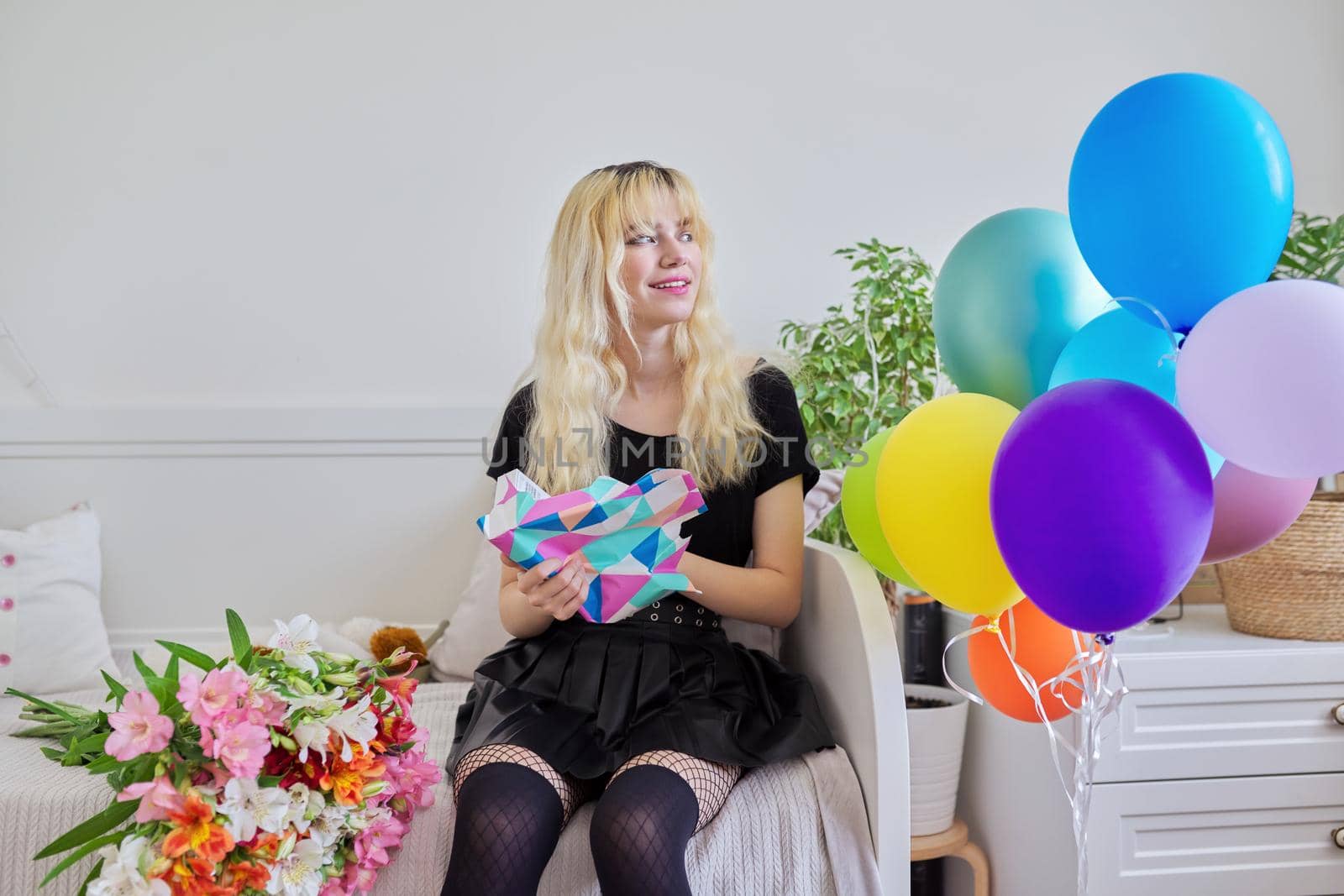 Birthday of teenage female 16, 17 years old. Hipster teenager at home with surprise gift in box, bouquet of flowers, colored balloons, sitting at home on bed. Teens, age, holiday concept