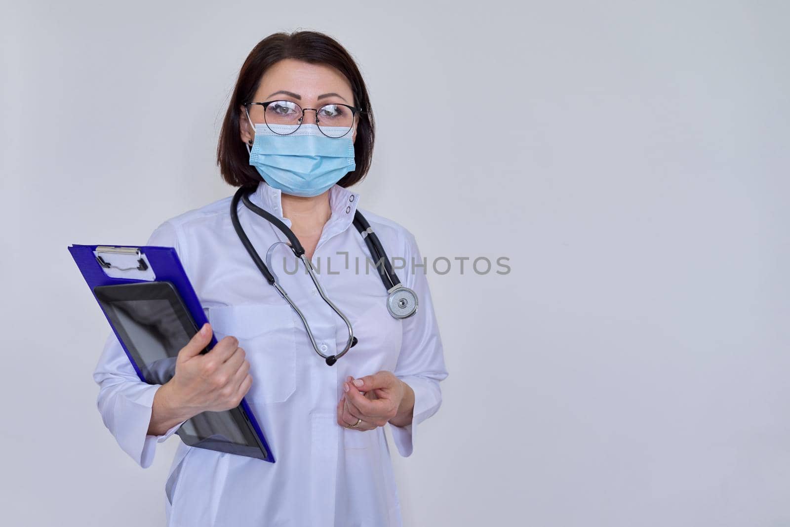 Portrait of female doctor in white uniform in protective medical face mask with stethoscope, with clipboard and digital tablet in her hands, woman looking at camera, on light background, copy space