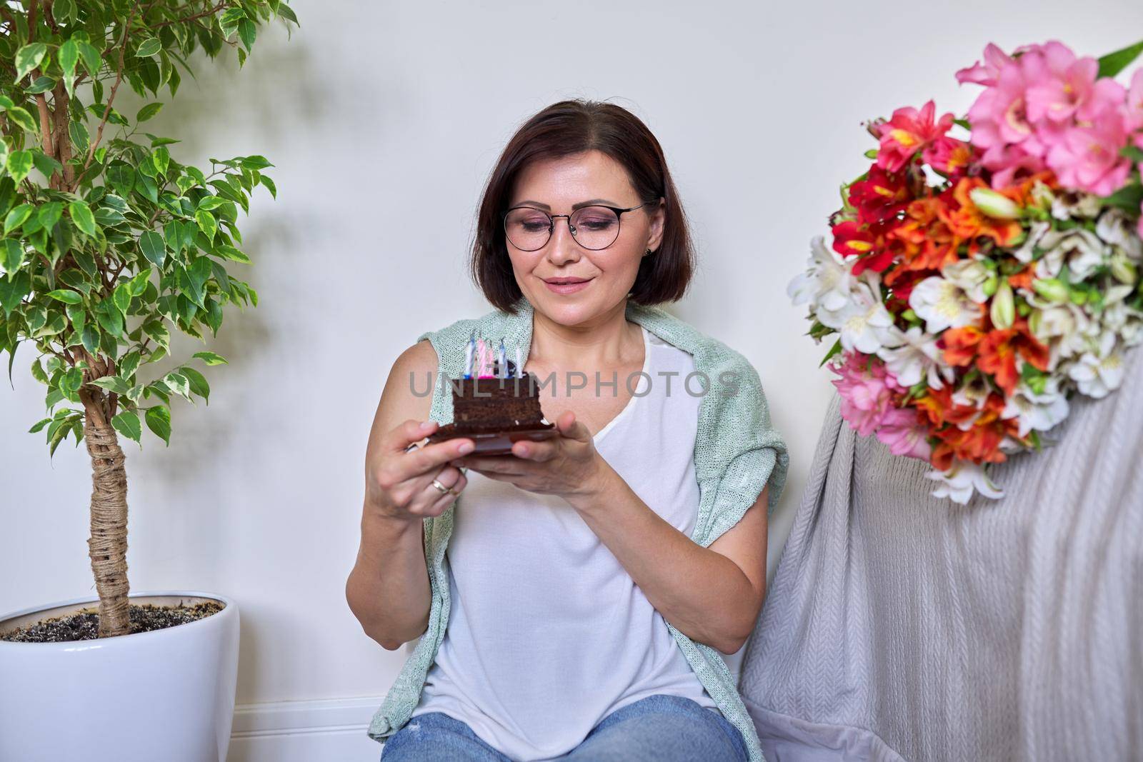 Middle-aged women with small birthday cake with candles. Happy female at home on the floor with bouquet of flowers. Age, date, aging, celebration, lifestyle, anniversary concept