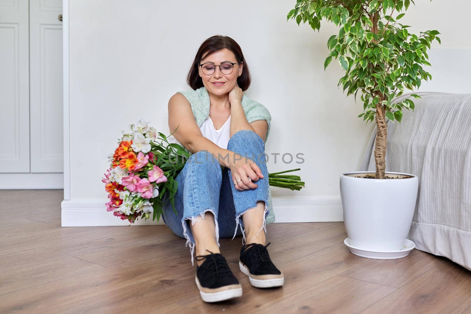 Smiling middle aged woman with bouquet of flowers sitting on the floor at home by VH-studio