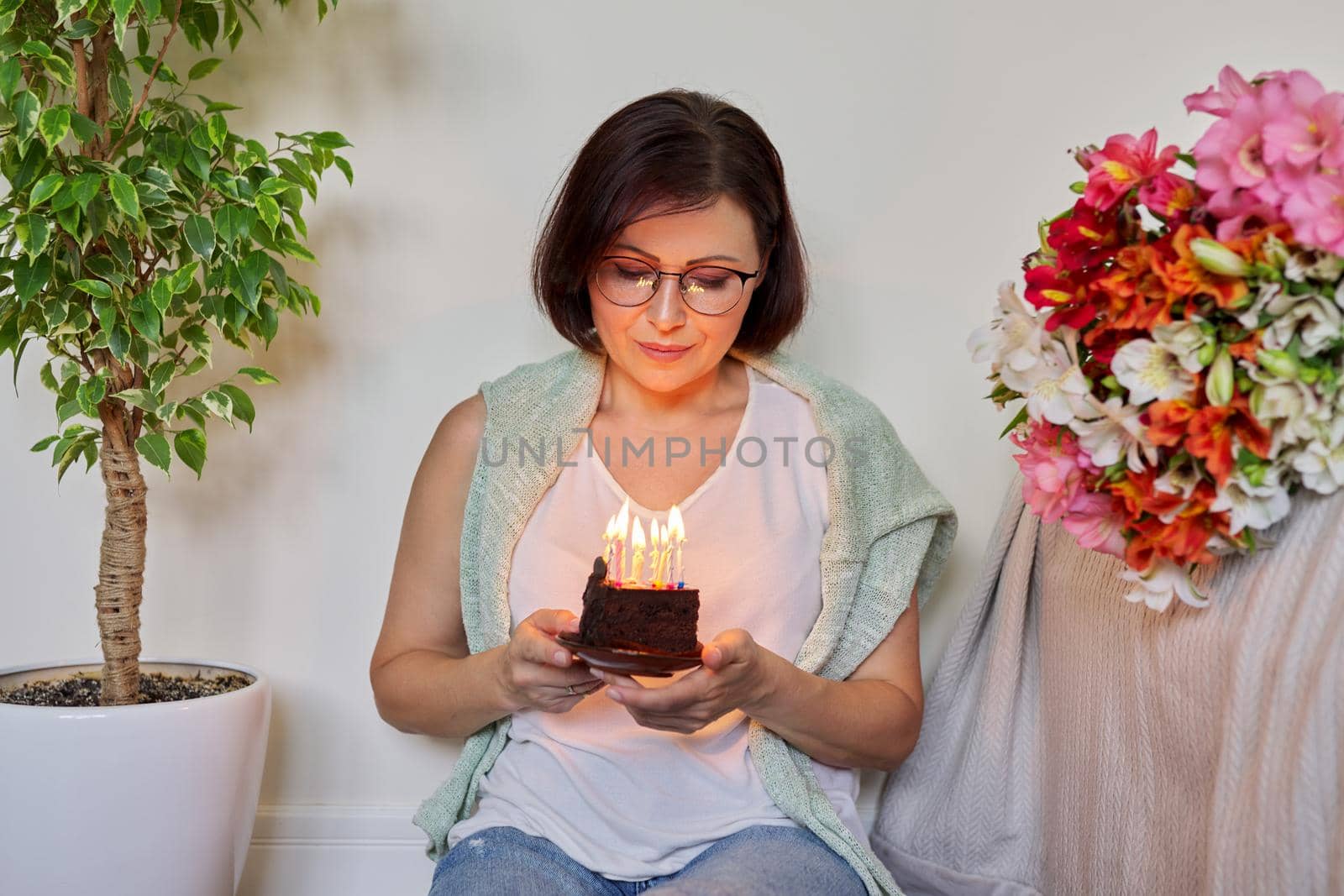 Birthday, celebration, anniversary, 45 years old woman with small cake with burning candles. Smiling middle aged woman with bouquet of flowers sitting on the floor at home.