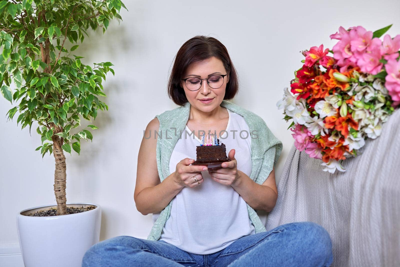 Birthday, celebration, anniversary, 45 years old woman with small cake with candles. Smiling middle aged woman with bouquet of flowers sitting on the floor at home.