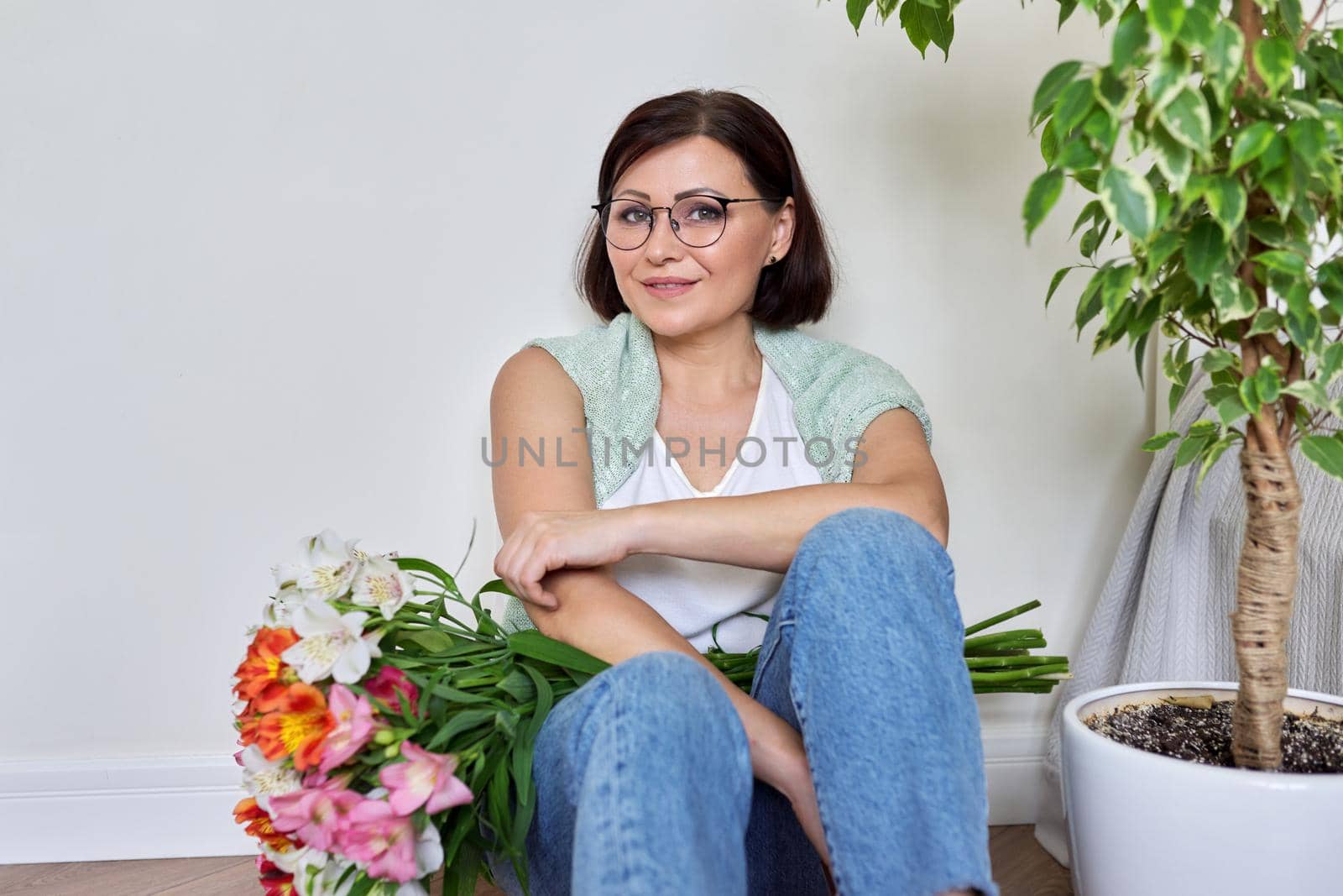 Smiling middle aged woman with bouquet of flowers sitting on the floor at home by VH-studio