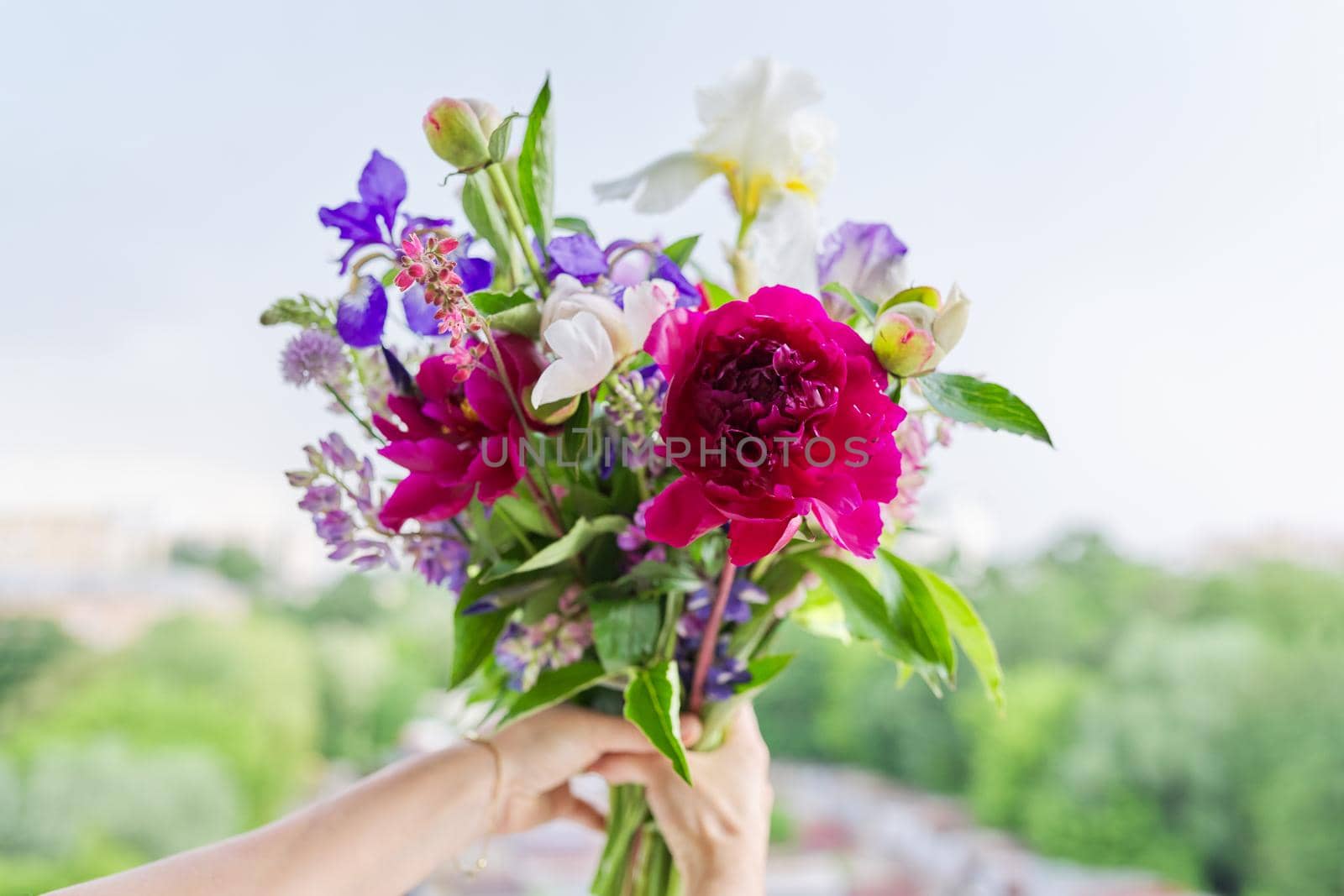 Close-up of bright bouquet of flowers in female hand. Fresh flowers and buds of peonies iris lupine in flower arrangement. Season spring summer, natural beauty, background open window blue sky