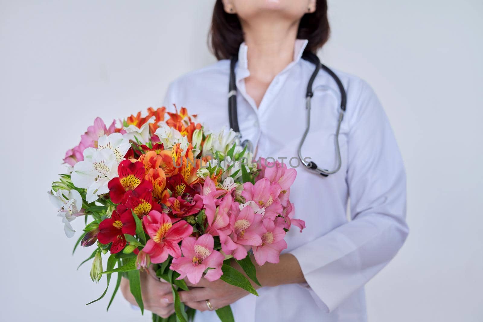 Close-up of bouquet of flowers in hands of female doctor. Medic, pharmacist in white uniform with stethoscope on light background. International, National Doctor's Day, Nurse's Day, Health Day