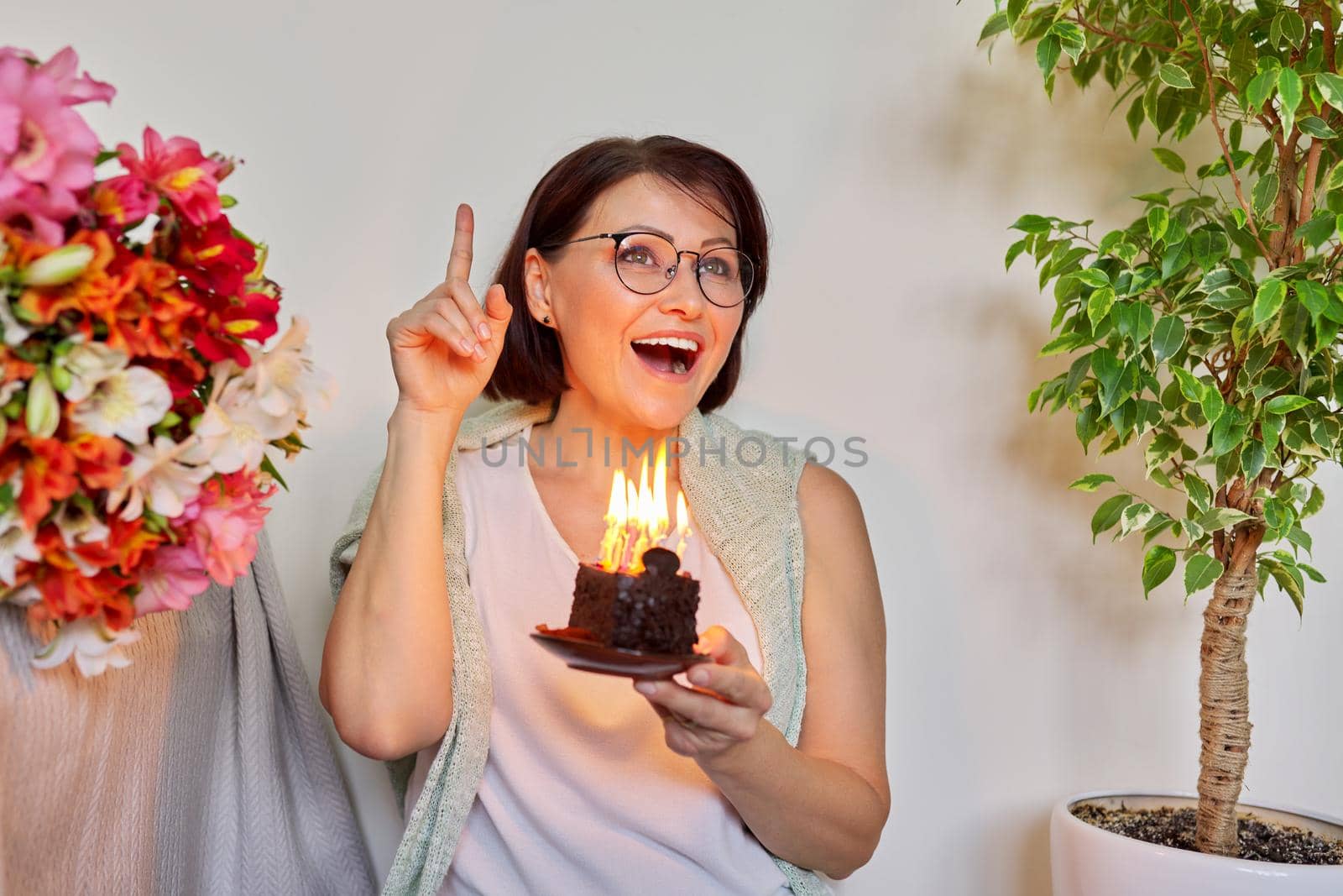 Mature woman with small birthday cake with burning candles. Happy 40s woman making wish before blowing out the candles. Age, birthday, anniversary, joy holiday happiness concept