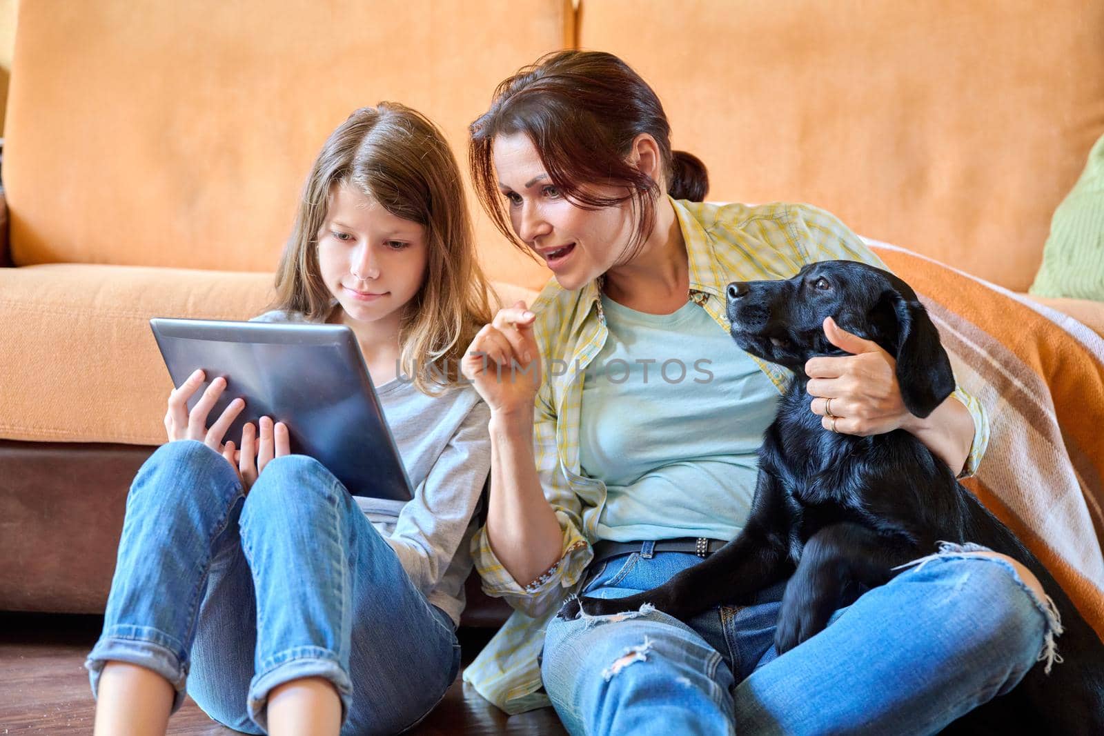 Family, lifestyle, communication, happiness, vacation together, pets, home concept. Mother daughter child and labrador puppy dog sitting together near sofa looking into digital tablet screen at home