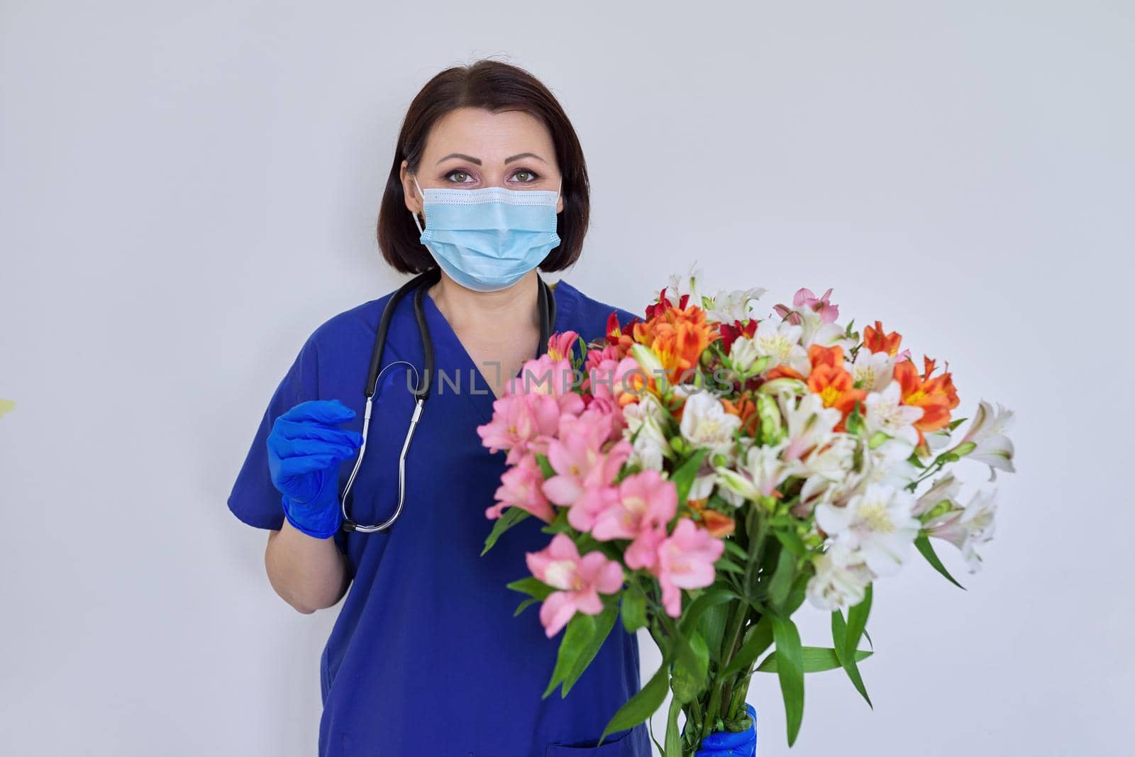 Female medic in blue uniform medical mask with bouquet of flowers on light background by VH-studio