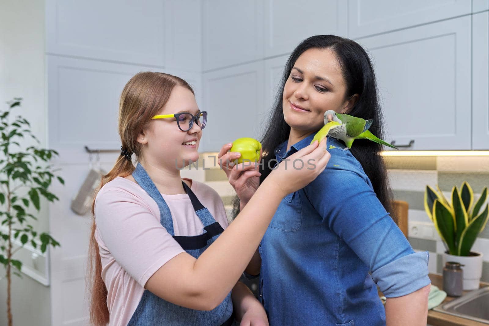 Friendly cheerful family, portrait of mom and teenage daughter, with green parrot. Girl feeding parrot pet sitting on shoulder with apple, kitchen house background