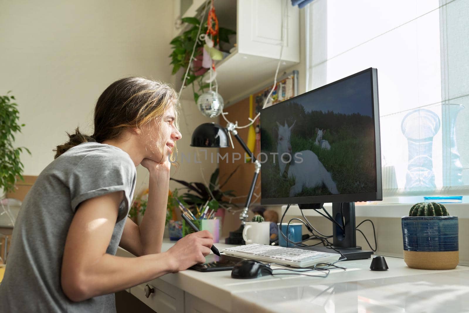 Teen guy sitting at table at home working on computer, using graphics tablet to work with images. Modern technologies, hobby of male teenager, freelance, youth concept