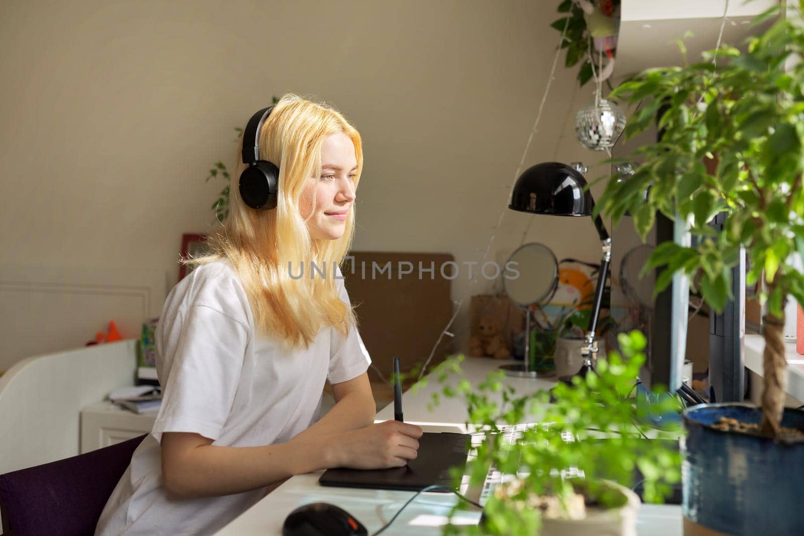 Girl teenager artist in headphones sitting at the table at home drawing on computer using graphics tablet. Hobbies and leisure of creative college student