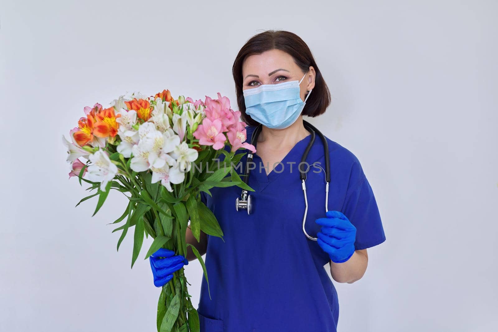 Doctor's Day, Nurse's Day. Female medic in blue uniform medical protective face mask gloves with stethoscope with large bouquet of flowers on light background