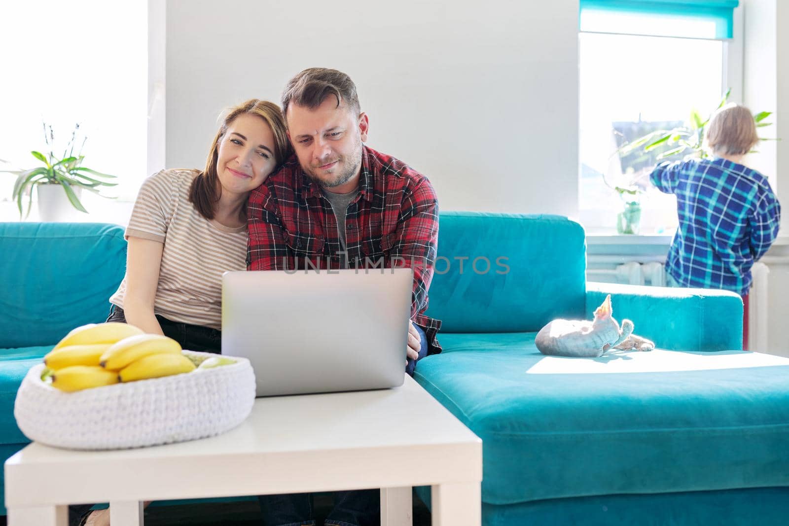 Smiling laughing positive couple 40 years old, husband and wife looking at laptop monitor sitting together at home on sofa in living room, child son near the window, real people life