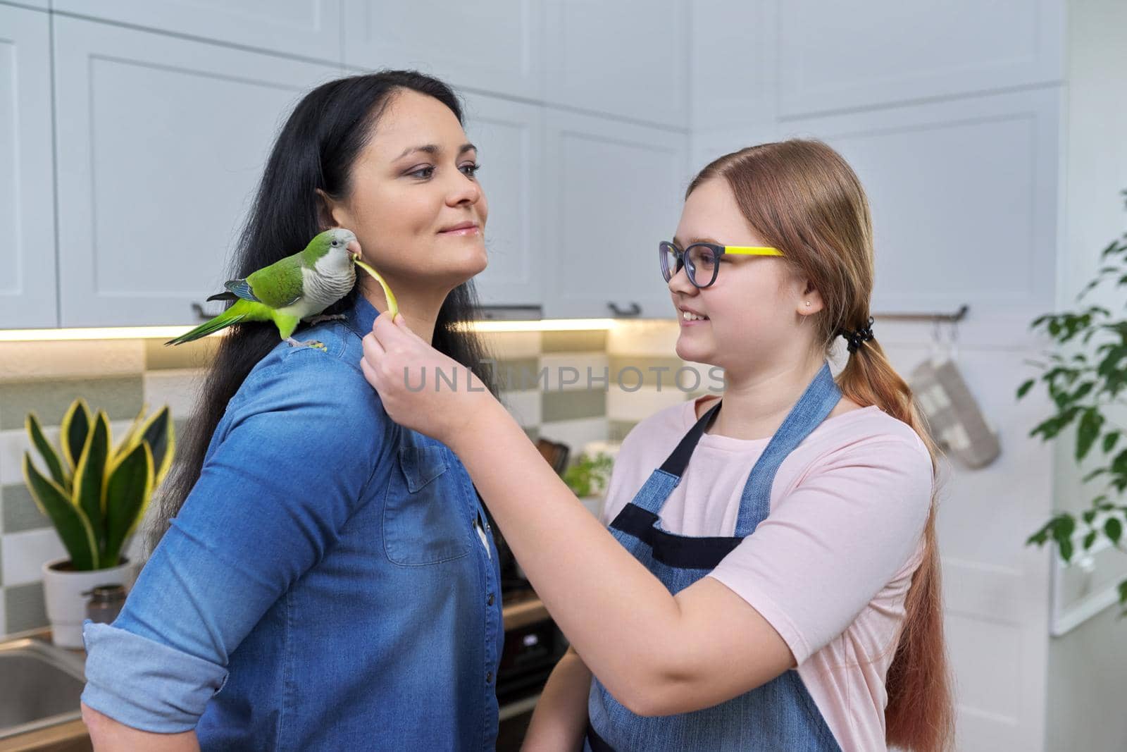 Friendly cheerful family, portrait of mom and teenage daughter, with green parrot. Girl feeding parrot pet sitting on shoulder with apple, kitchen house background