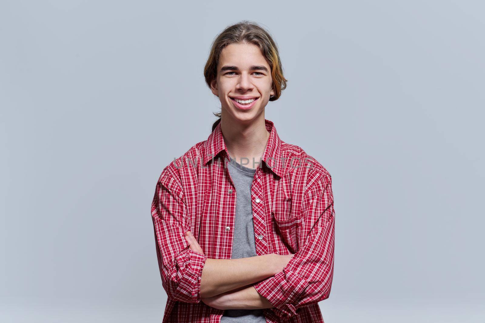 Portrait of smiling guy teenager with crossed arms, hipster teen male 15, 16 years old with long hair looking at camera on light background