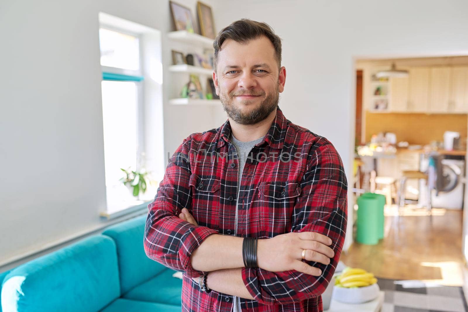Portrait of confident middle aged man, smiling male in plaid casual shirt looking at camera with crossed arms, home living room interior background