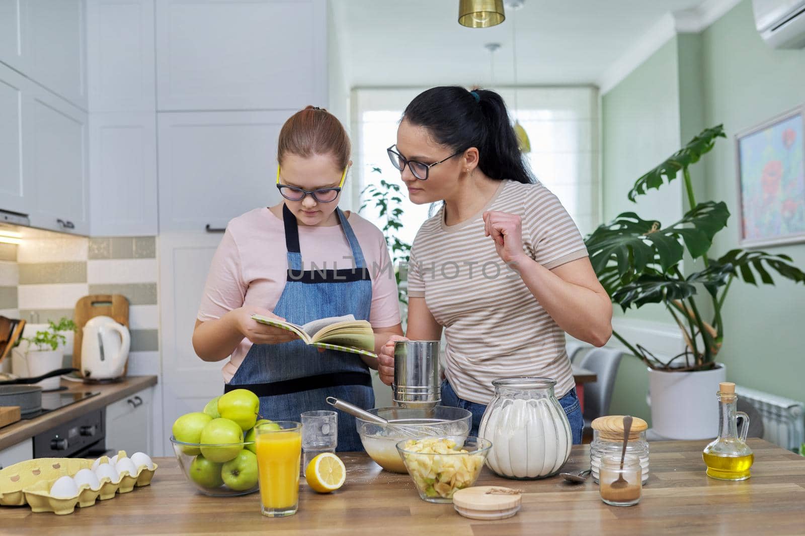 Mom and teenage daughter preparing apple pie in kitchen together, looking at recipe book, talking and laughing. Communication between teenager and parent, lifestyle, cooking at home concept