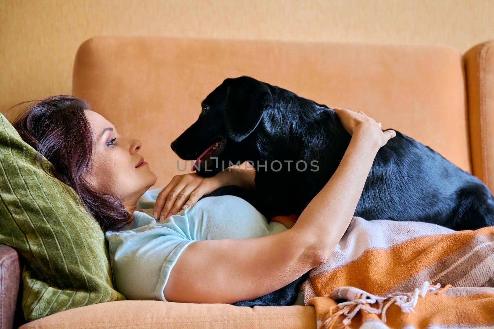 Relaxed middle aged woman lying on sofa under blanket and petting black labrador puppy dog. Lifestyle, love, care, pets, 40s people concept