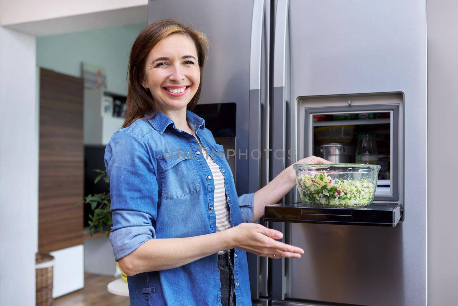 Woman with bowl of vegetable salad from refrigerator. Kitchen interior, open modern chrome refrigerator door background. Eating at home, lifestyle, household, dieting, healthly food concept