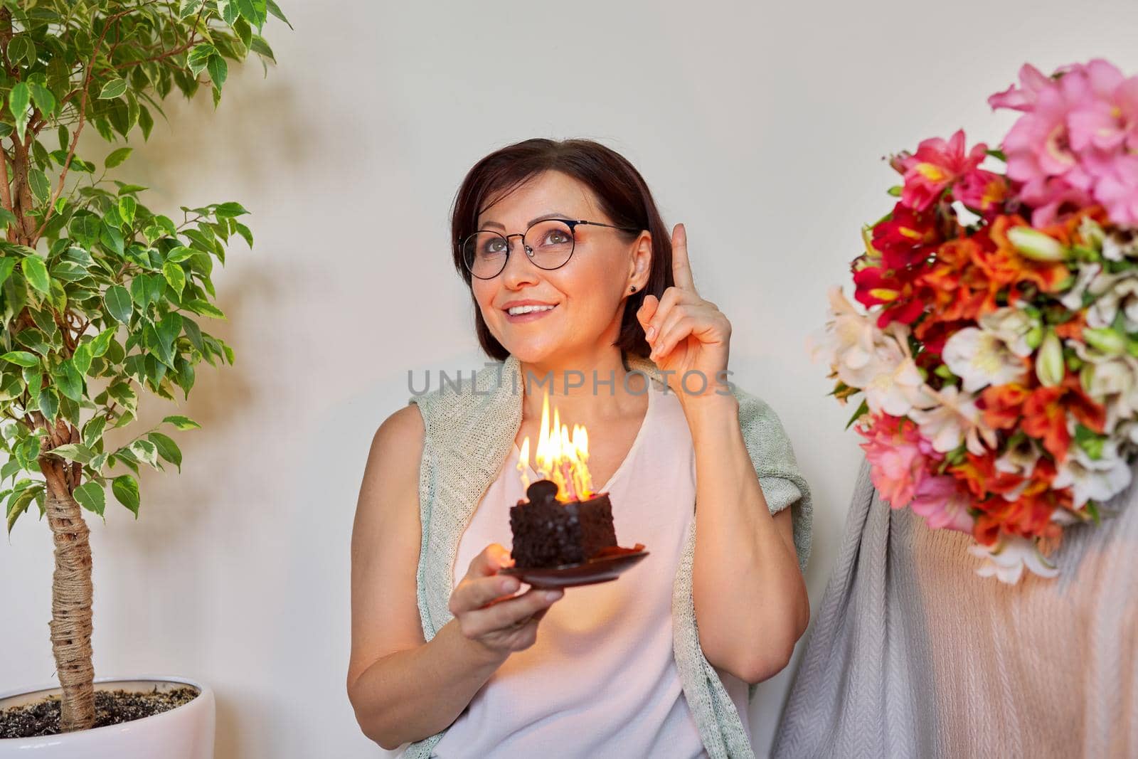 Mature woman with small birthday cake with burning candles. Happy 40s woman making wish before blowing out the candles. Age, birthday, anniversary, joy holiday happiness concept