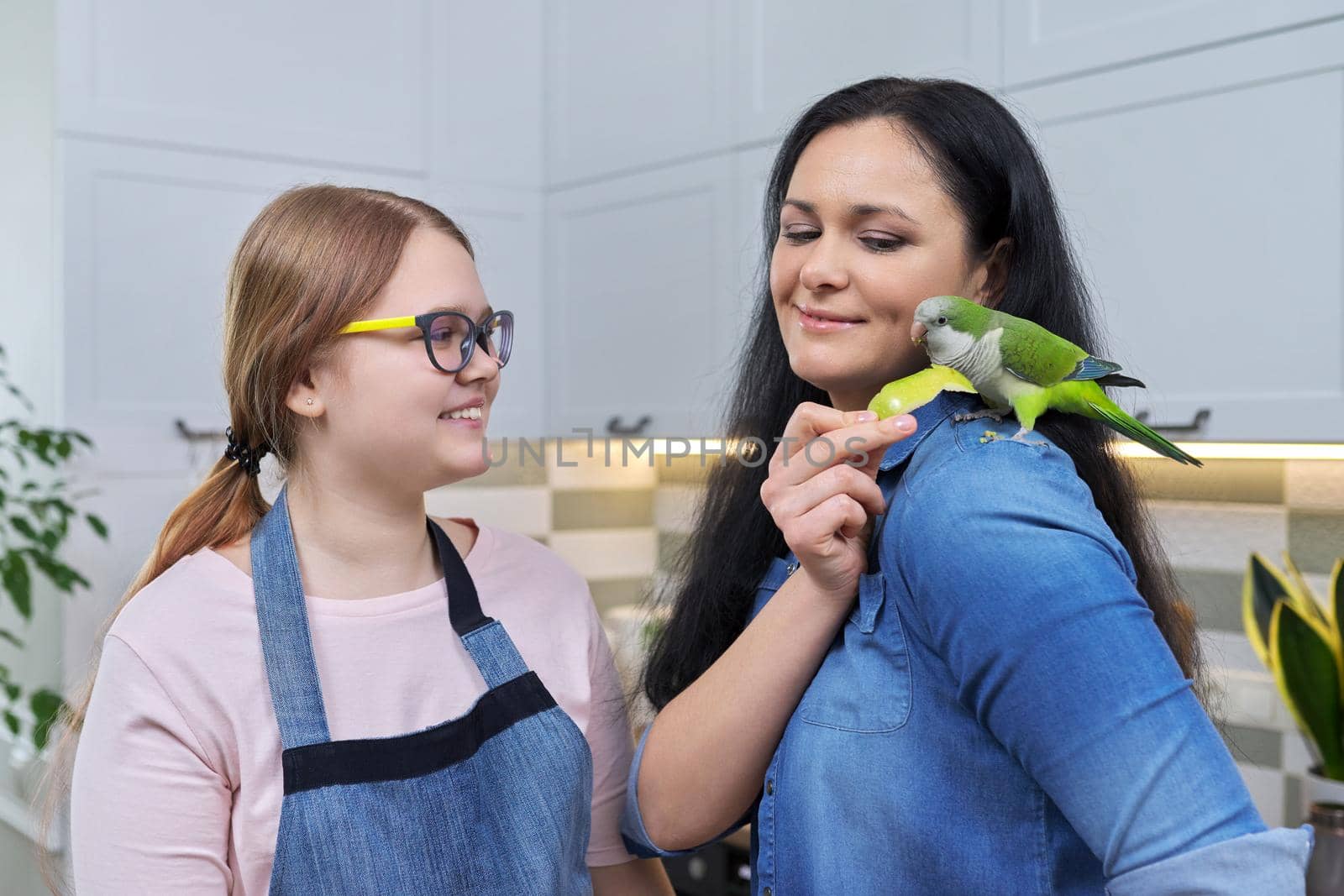 Friendly cheerful family, portrait of mom and teenage daughter, with green parrot. Woman feeding parrot pet sitting on shoulder with apple, kitchen house background