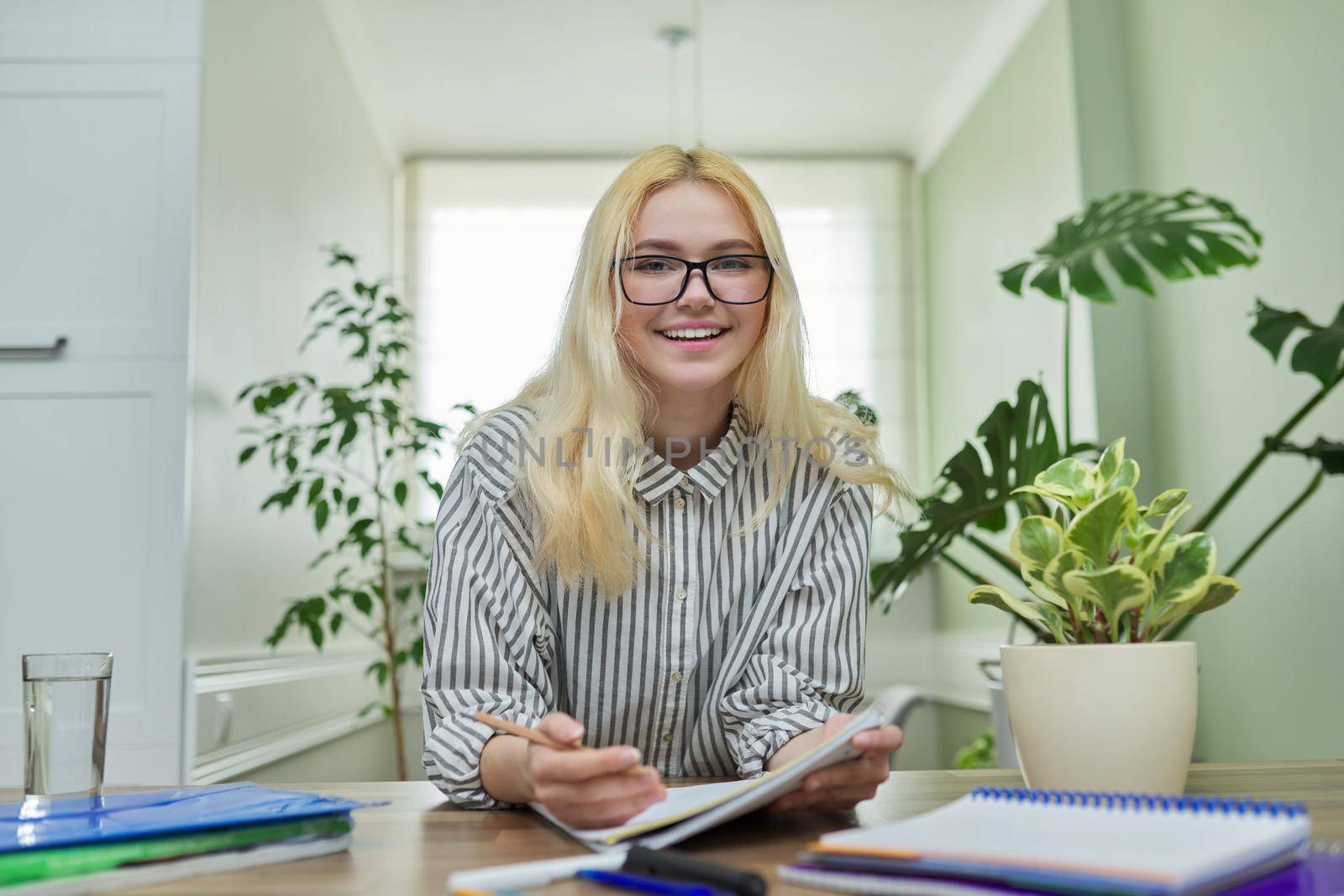 Portrait of teenager female student looking at camera, talking smiling, sitting at home at the table with notebooks textbooks. Looks in webcam, online education, video lesson, conference, e-learning