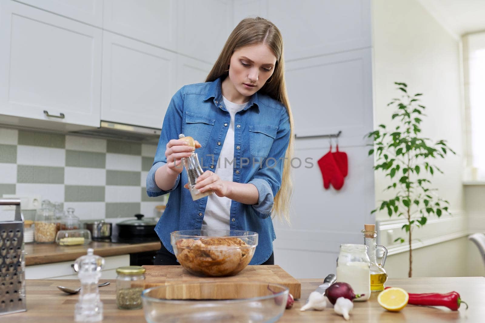 Young woman cooking chicken, marinating with spices black pepper salt, at home in kitchen by VH-studio