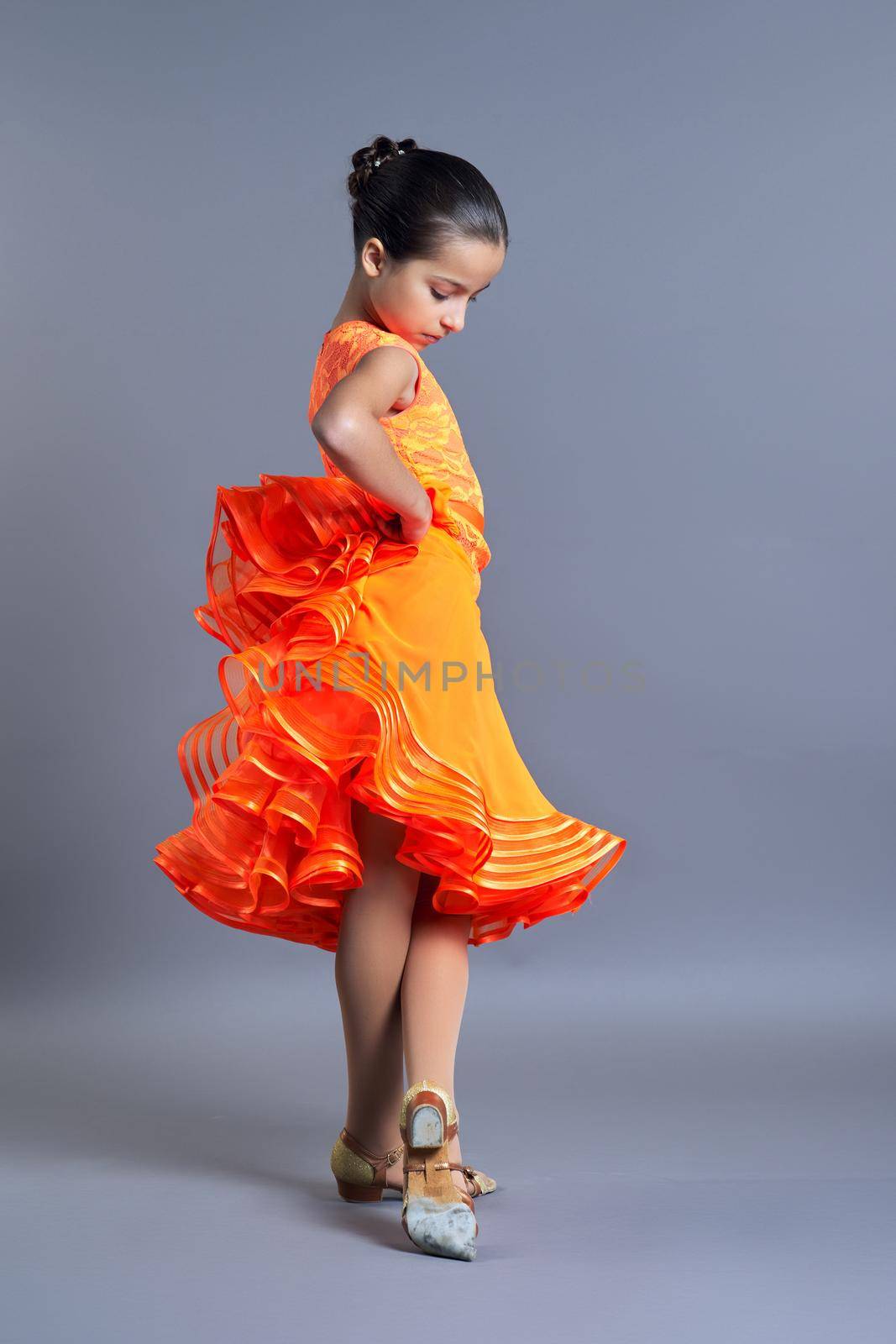 Child girl in an orange sports dress posing in dance movement on gray background by VH-studio