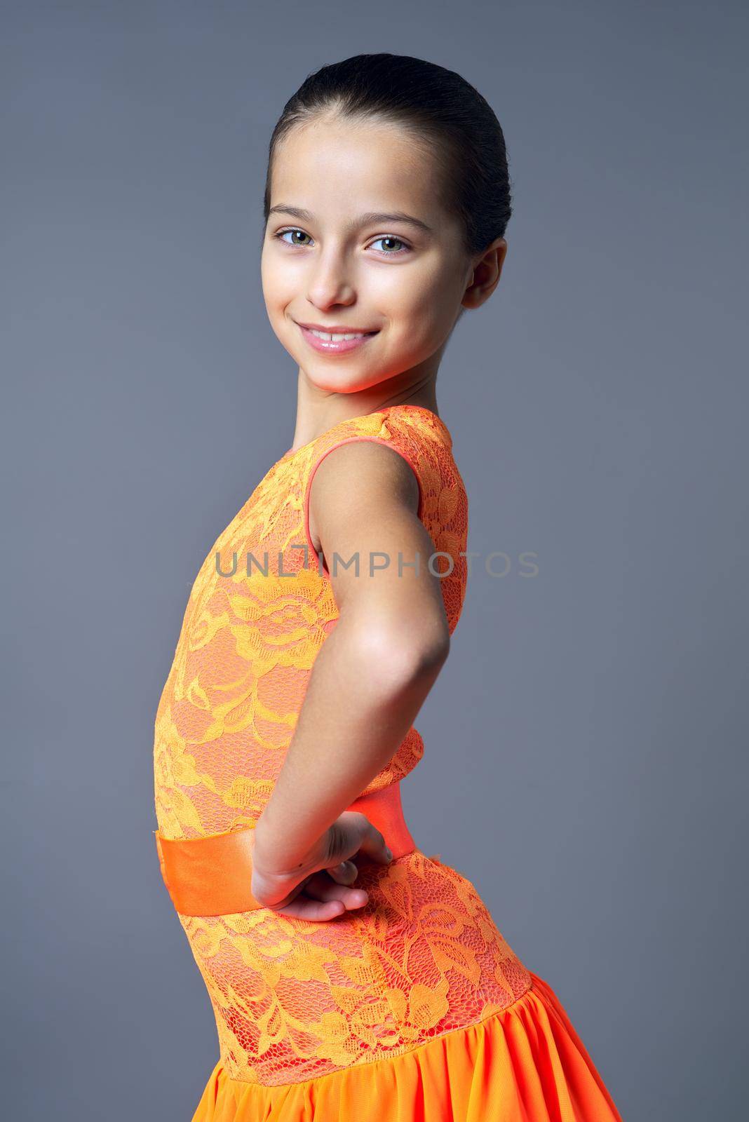 Portrait of smiling child girl in orange sports dress posing looking at camera on gray studio background, sport dances ballroom and latin american for children and teenagers