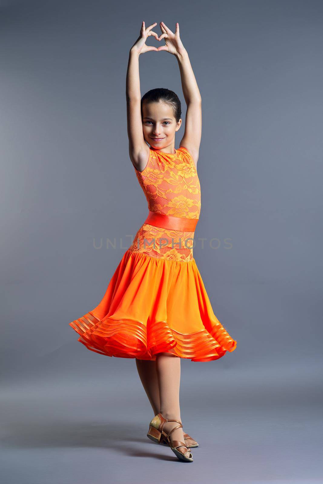 Kids sport dancing, kid girl in orange sport dress posing in dance pose looking at camera on gray studio background.