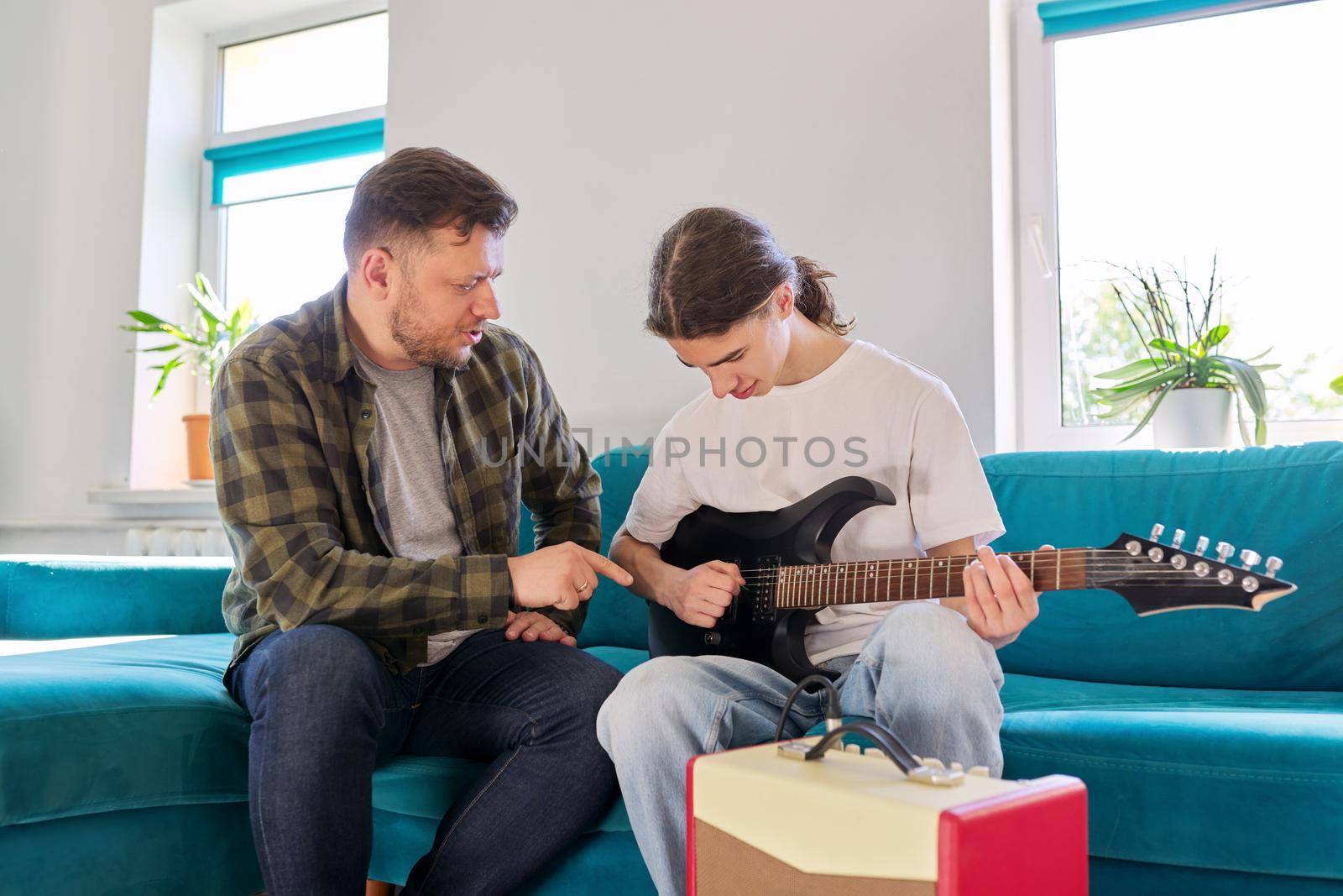 Father teaches his teenage son to play the electric guitar. Family sitting together at home in living room on couch. Parent-child relationship, lifestyle, creativity, music, teenagers concept