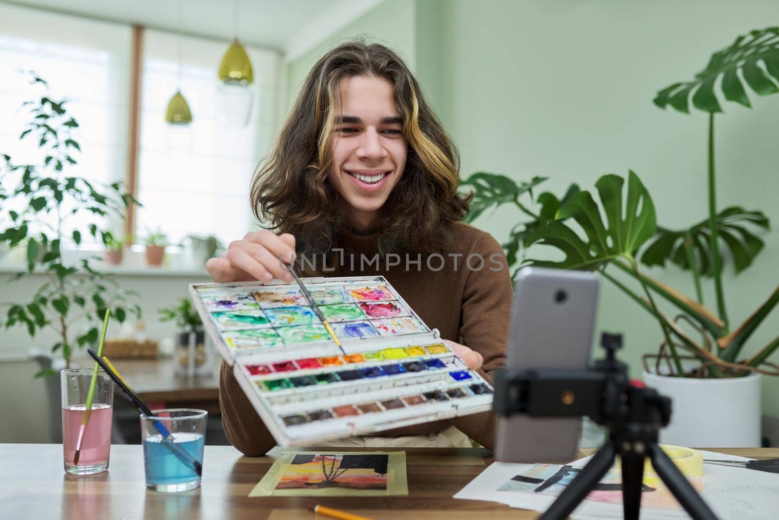 Online learning, drawing, e-education. Teenage guy drawing with watercolors, watching video class on smartphone, sitting at table at home. Creativity, technology, hobby, teens concept