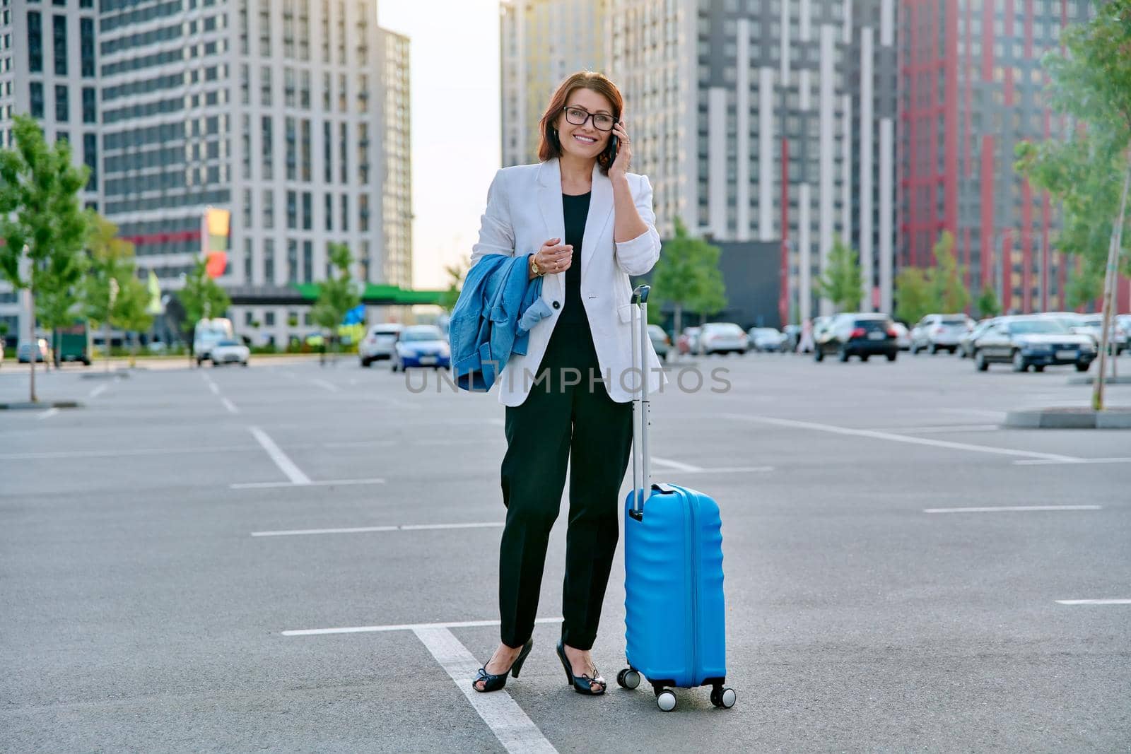 Middle aged businesswoman with suitcase talking on mobile phone in outdoor parking, urban style, city architecture background. Business travel trip, business people of mature age, success, modern city