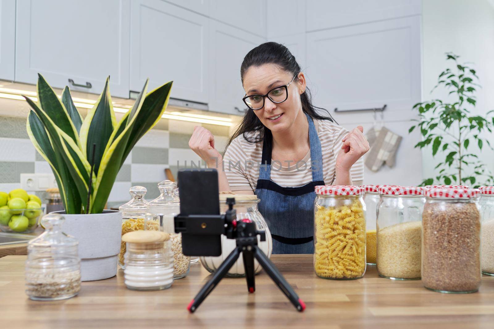 Online broadcast, blog about food, woman in an apron with jars of cereals noodles flour, looking at smartphone camera, telling and showing about food storage, organizing kitchen