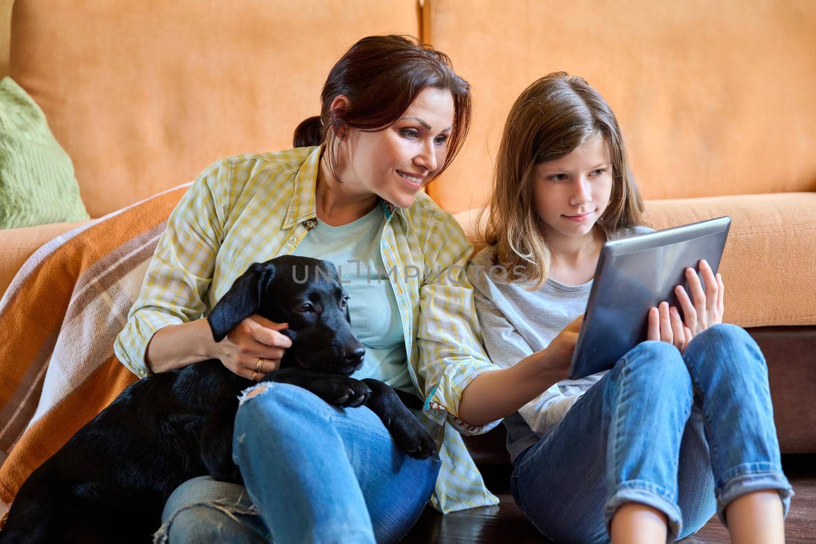 Family, lifestyle, communication, happiness, vacation together, pets, home concept. Mother daughter child and labrador puppy dog sitting together near sofa looking into digital tablet screen at home