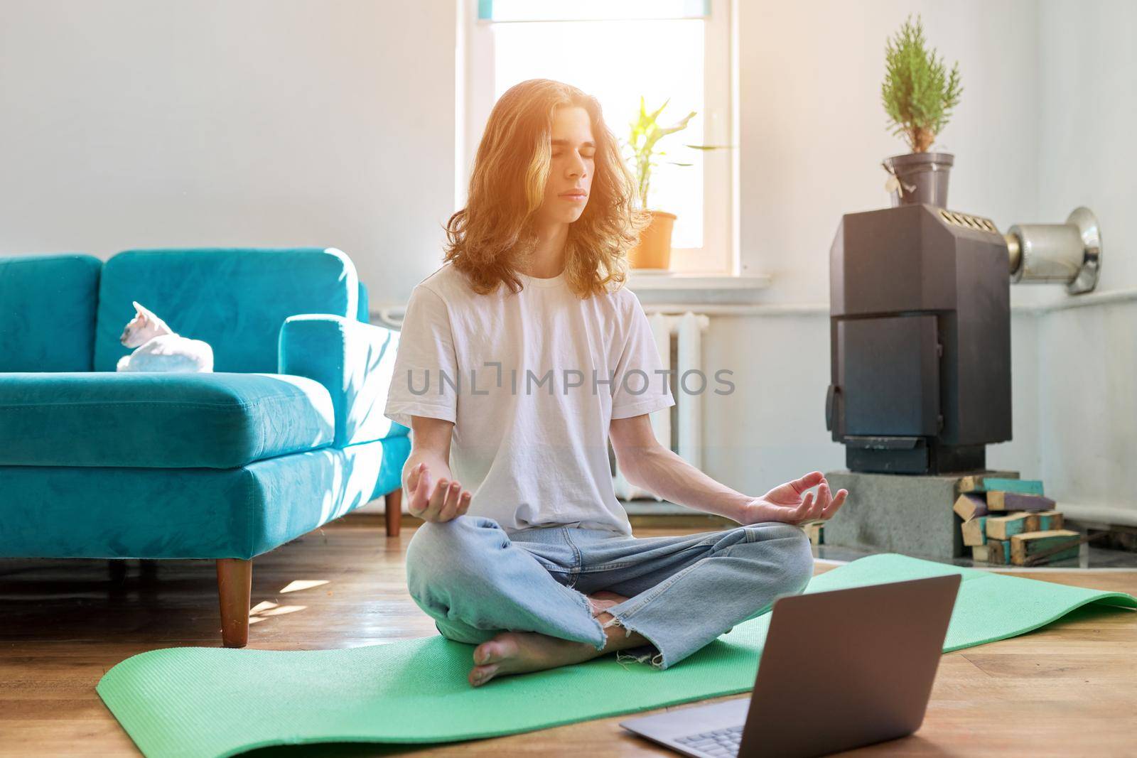 Guy teenager sitting in lotus position on yoga mat at home on floor with laptop by VH-studio