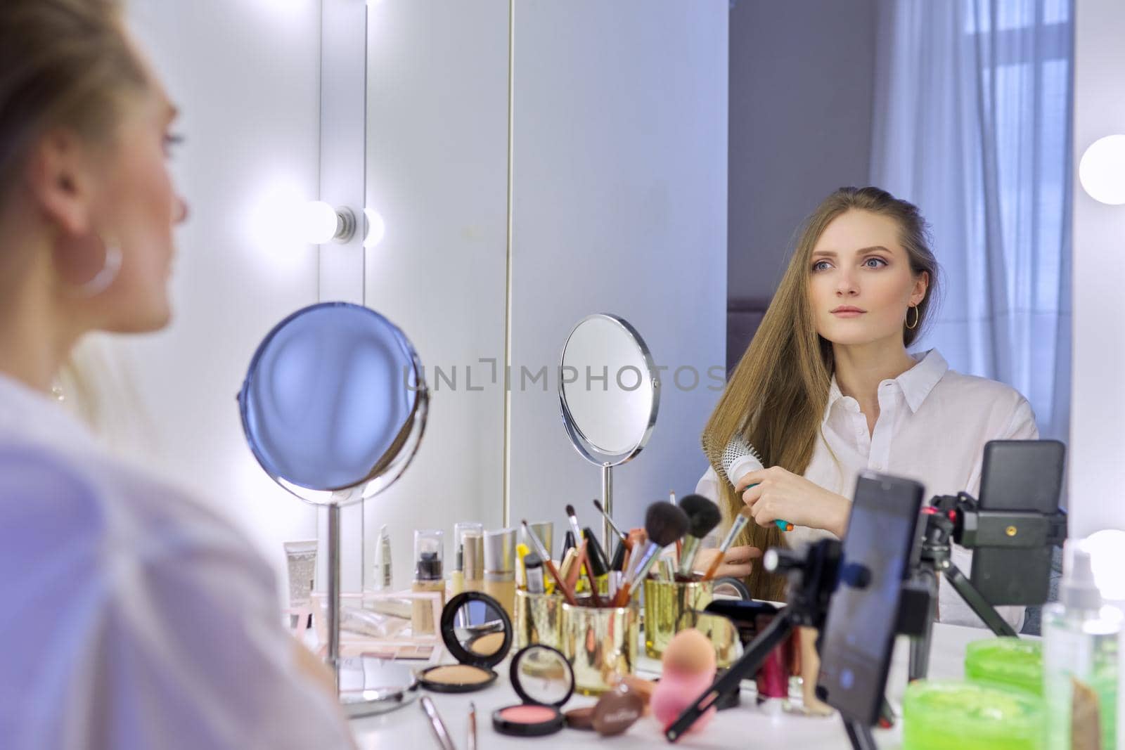 Young beautiful woman combing hair in front of mirror, blonde with long healthy straight hair. Beauty blogger recording video on smartphone, online course, remote training in makeup and hair styling