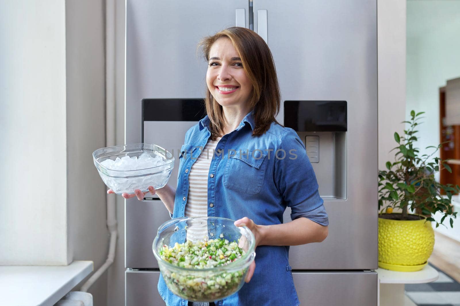 Smiling woman with bowl of ice and plate of vegetable salad in her hands, modern chrome large refrigerator in kitchen. Eating at home, lifestyle, household, dieting, healthly food concept