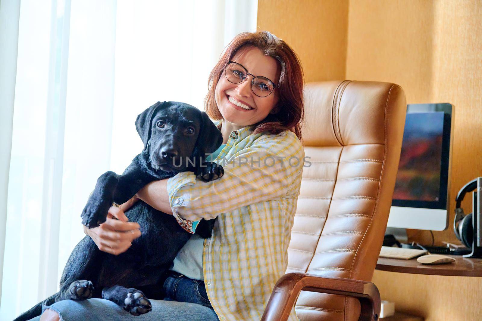 Portrait of middle aged woman and black labrador puppy dog. The owner holding young pet in her arms, in home interior. Lifestyle, love, pets, 40s people concept
