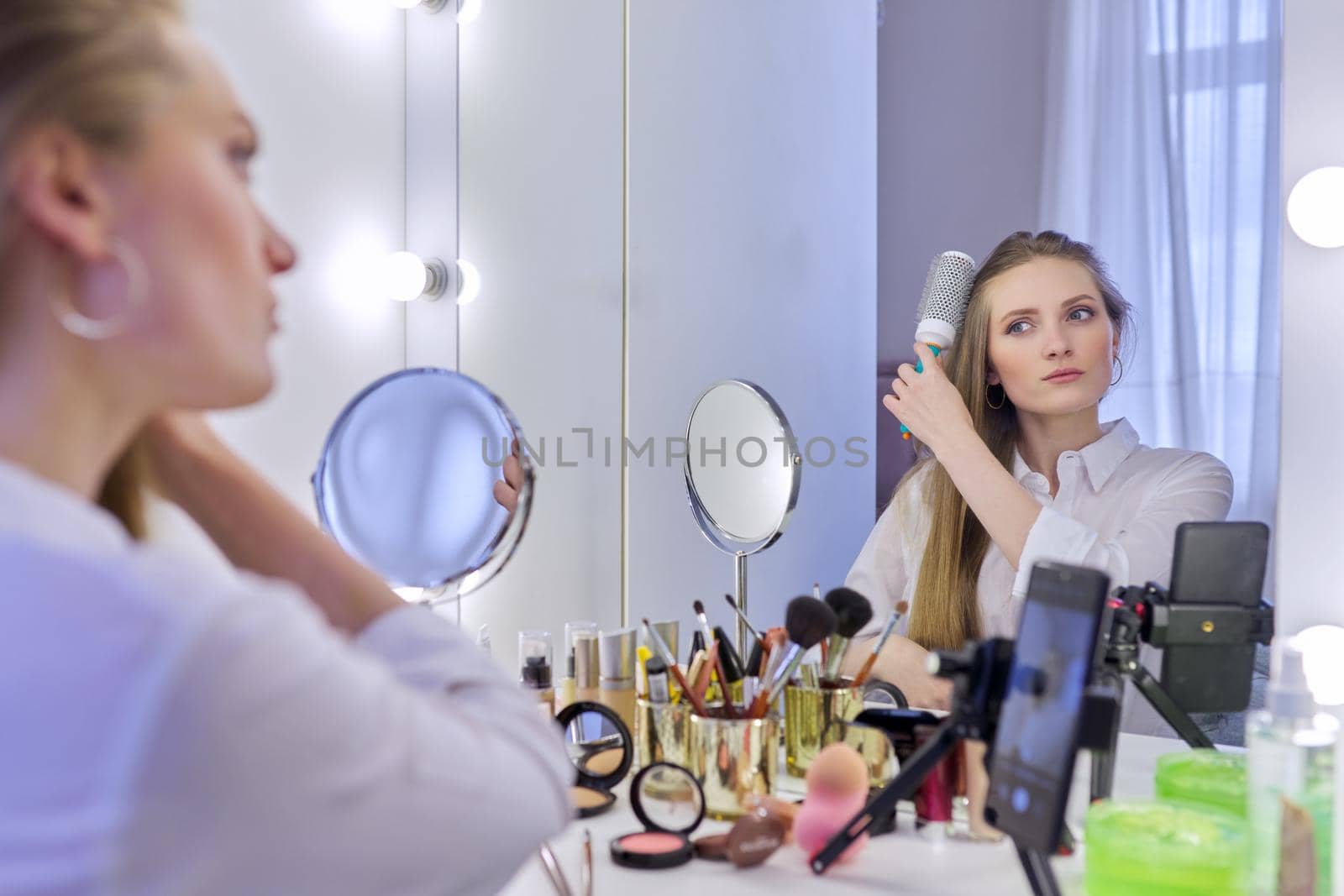 Young beautiful woman combing hair in front of mirror, blonde with long healthy straight hair. Beauty blogger recording video on smartphone, online course, remote training in makeup and hair styling