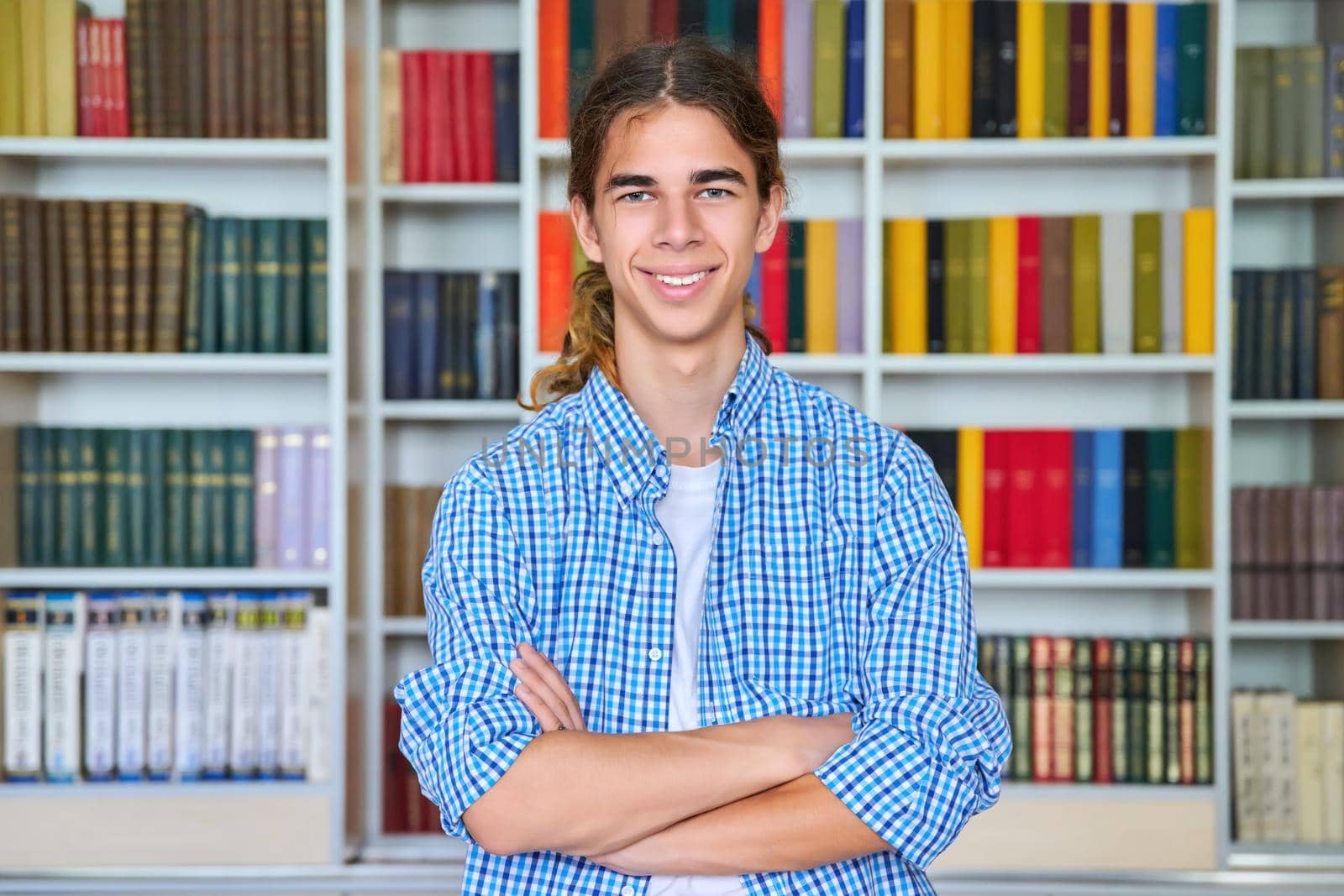 Single portrait of smiling confident male student teenager with crossed arms looking at camera in the library. School, education, knowledge, adolescence concept