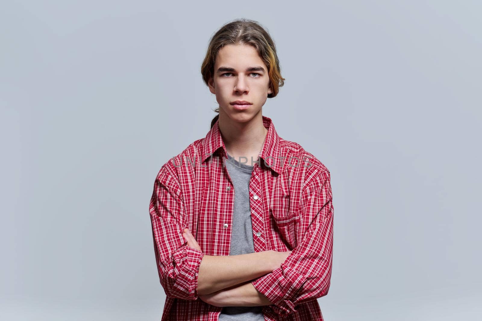 Portrait of serious guy teenager with crossed arms, hipster teen male 15, 16 years old with long hair looking at camera on light background