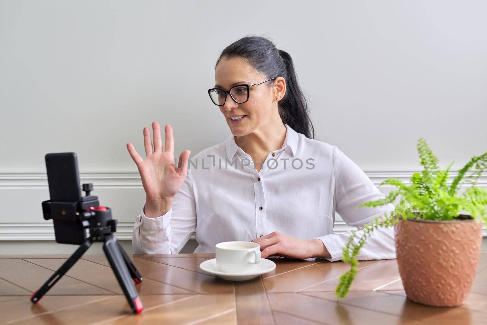 Young woman blogger vlogger recording broadcast at home using smartphone on tripod to communicate with followers. Female at the table with cup of coffee