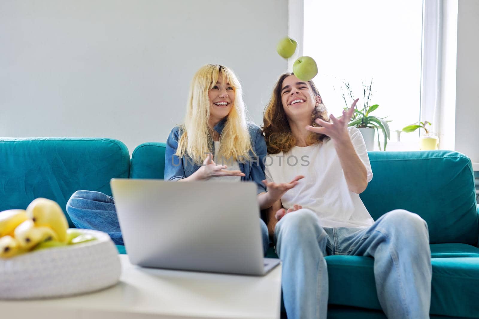 Laughing couple of teenagers having fun, sitting on couch, looking at laptop screen, with green apples in hands. Teens, youth, friendship, lifestyle, leisure concept
