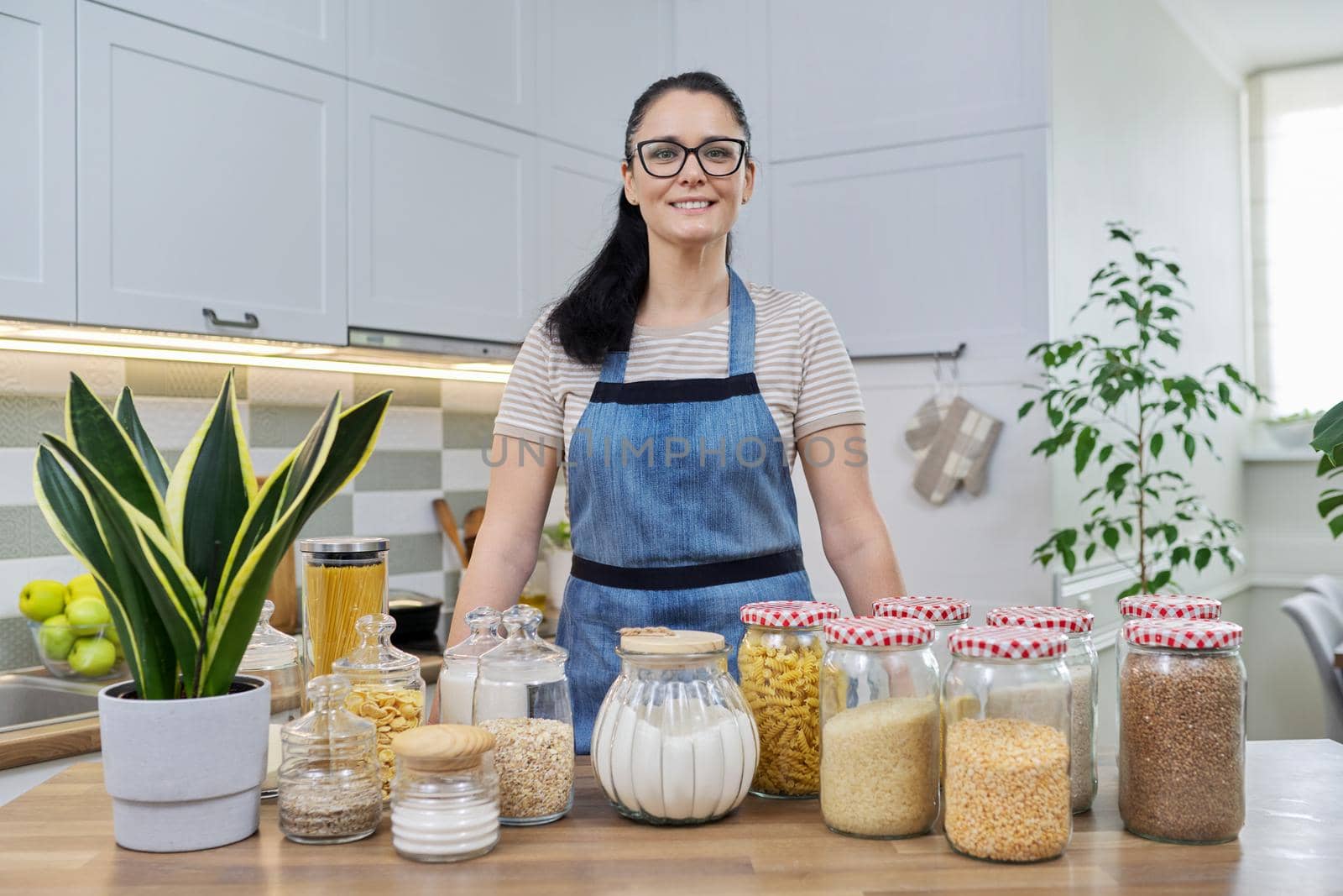 Portrait of smiling woman housewife in an apron in kitchen. Jars of cereals on the table, organizing and storing food