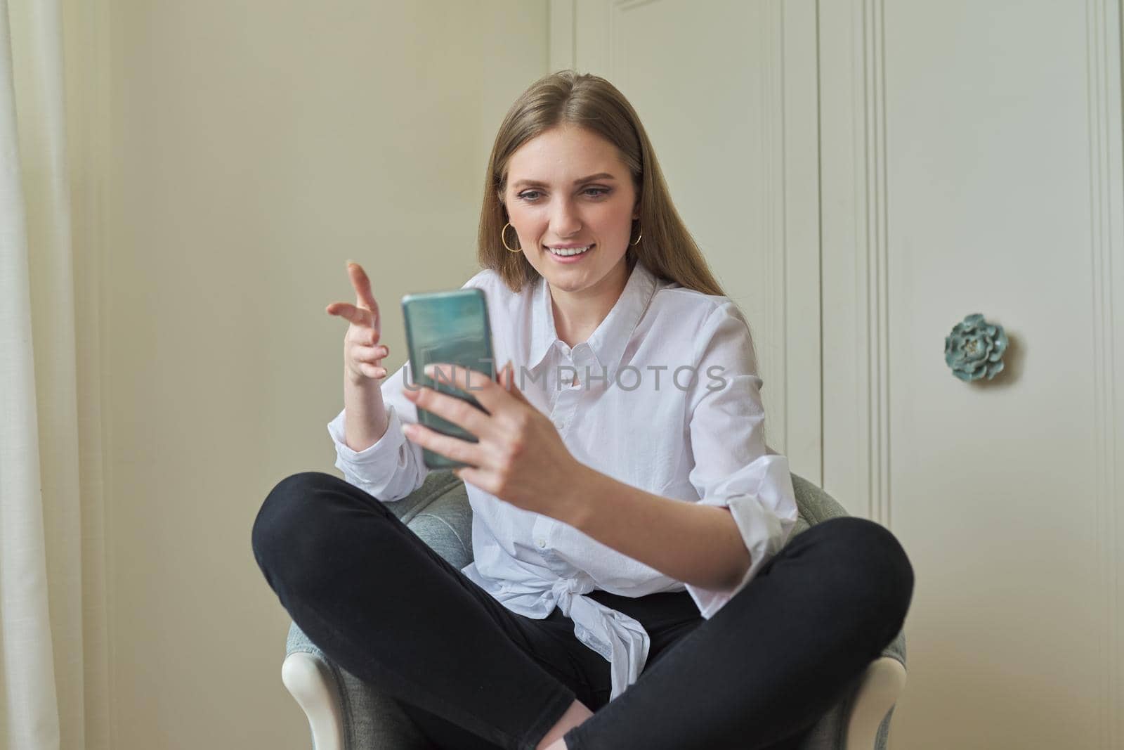 Emotional laughing young beautiful woman looking at smartphone screen, relaxing sitting at home in chair. Video call, emotional communication, joy, happiness, surprise, technology in everyday life