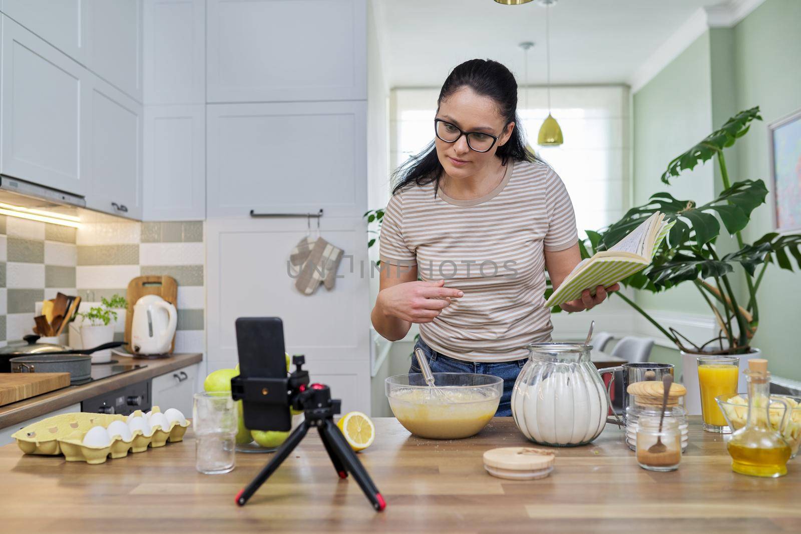 Woman preparing apple pie at home in kitchen with recipe book in her hand, looking in smartphone webcam, talking online using video call. Female food blogger recording video recipe